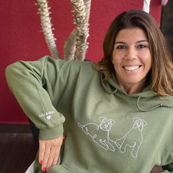 Smiling woman wearing her green custom embroidered hoodie with line art of her two dogs. The name of her dogs and a heart is engraved in the sleeve.