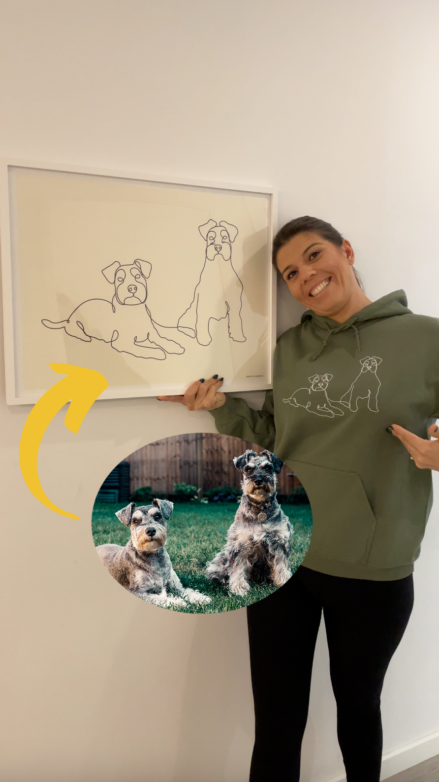 Woman holding a framed poster of line art of her two dogs. Woman wearing a green custom embroidered hoodie with line art of her two dogs.