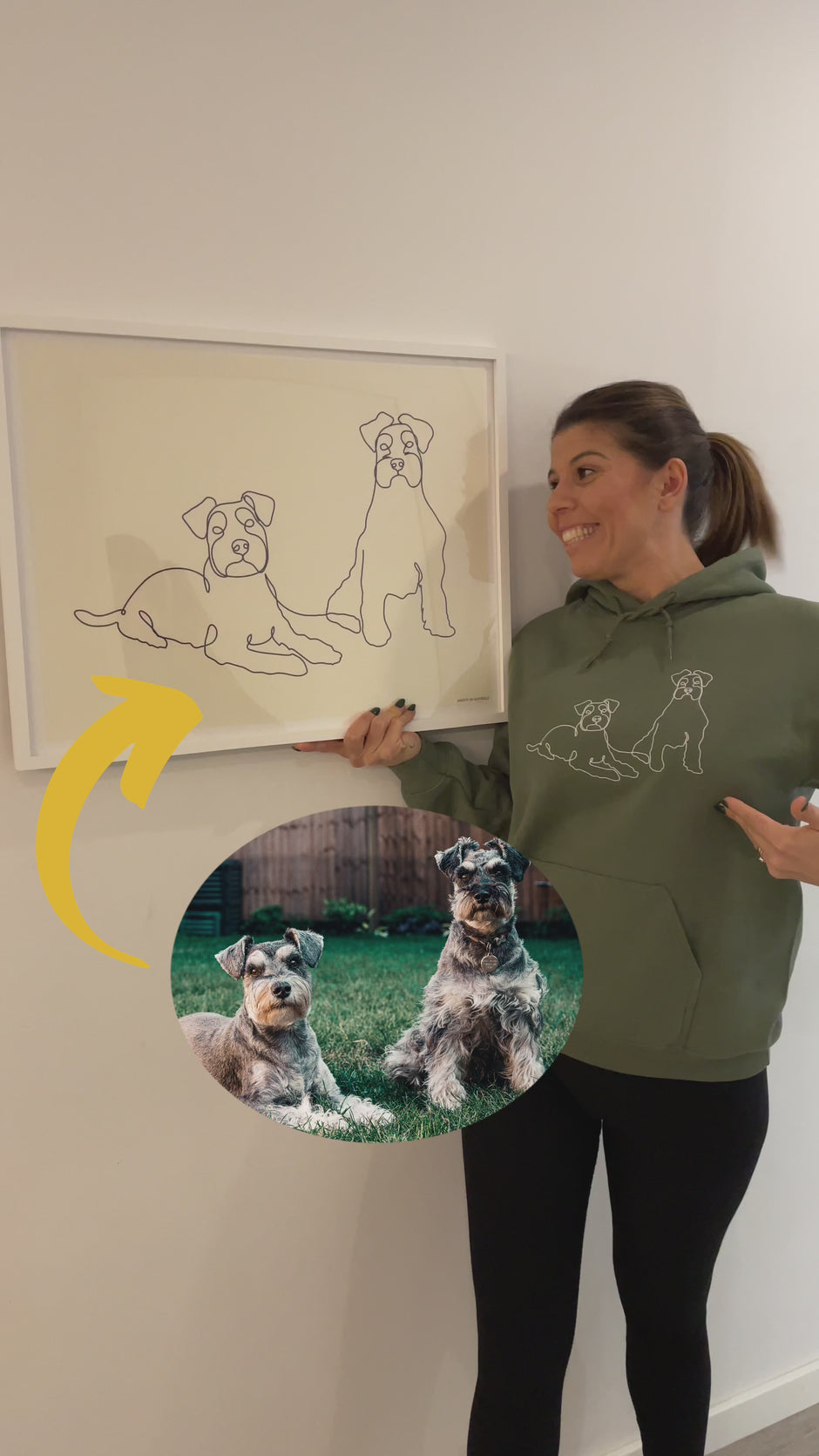 Woman holding a framed poster of line art of her two dogs. Woman wearing a green custom embroidered hoodie with line art of her two dogs.