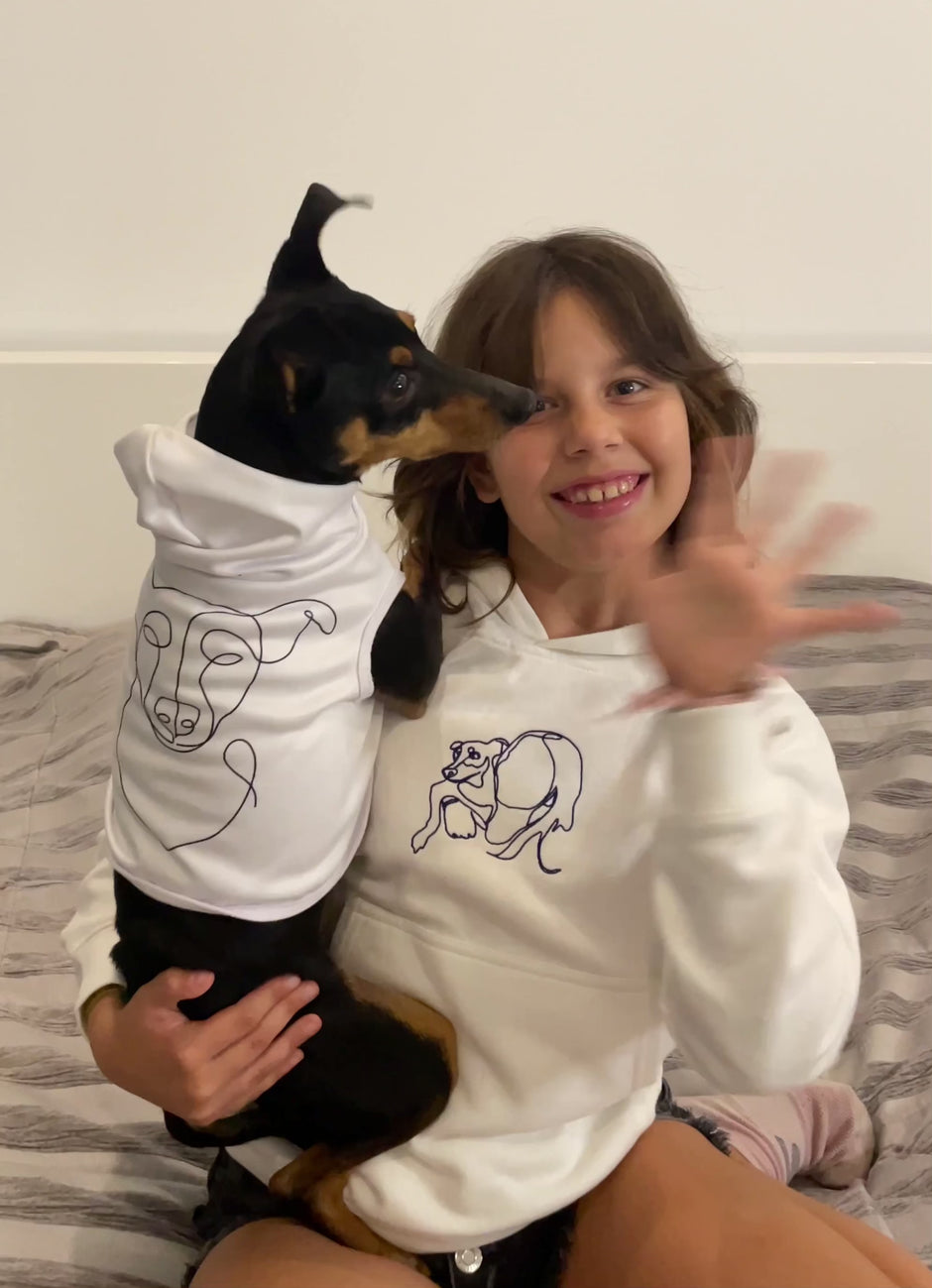 Happy kid holding her dog. The kid has a custom embroidered hoodie with line art of herself holding her dog. The dog has a custom hoodie with line art of his face. The kid is waving at the camera, smiling. The dog is looking around.